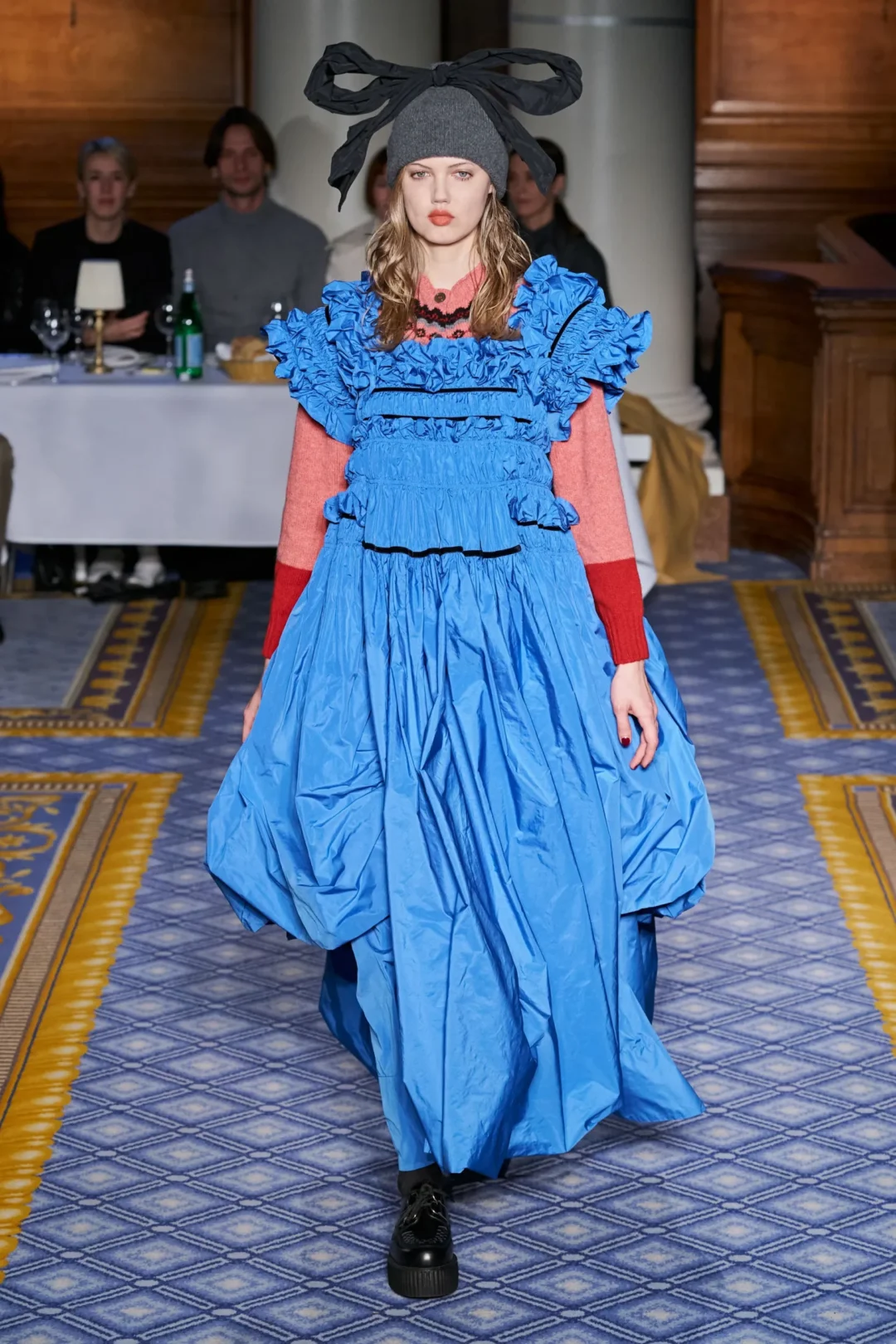 A model walking the catwalk, wearing a blue dress and a hat with a bow on it.